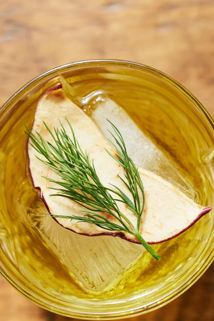 Golden cocktail in glass, garnished with dried apple and dill sprig.