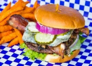 Gourmet burger with cheese, pickles, and sweet potato fries on checkered paper.