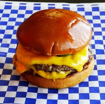 Close-up cheeseburger with melted cheese and a pickle on blue checkerboard paper.