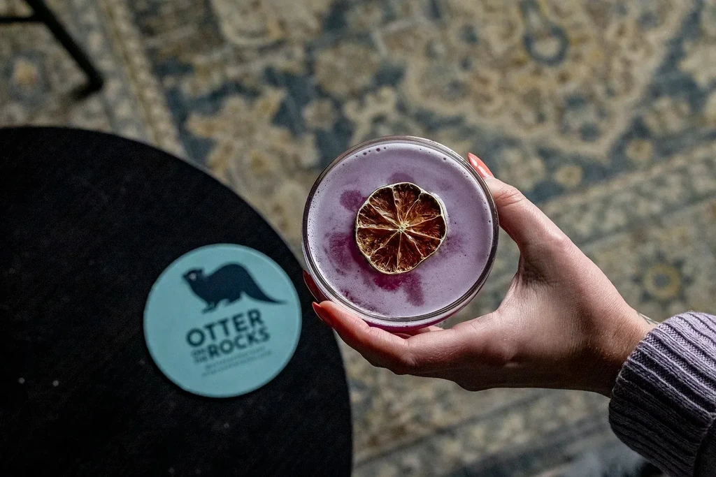 Hand holding purple cocktail with citrus garnish, "Otter on the Rocks" coaster.