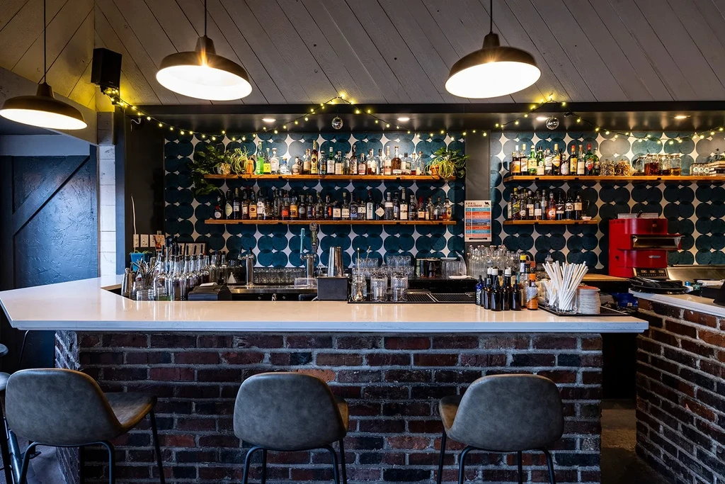 Stylish bar with white counter, liquor shelves, teal wallpaper, and warm lighting.
