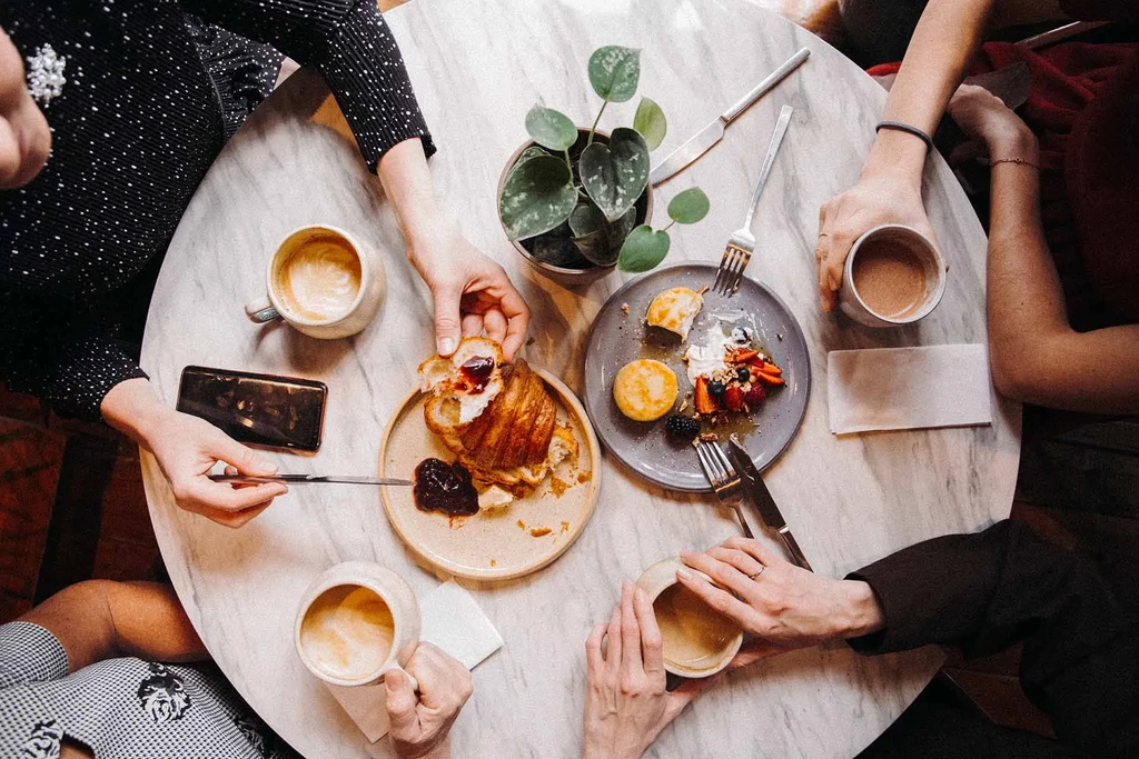 Overhead of hands dining on breakfast: croissant, pancakes, coffee.