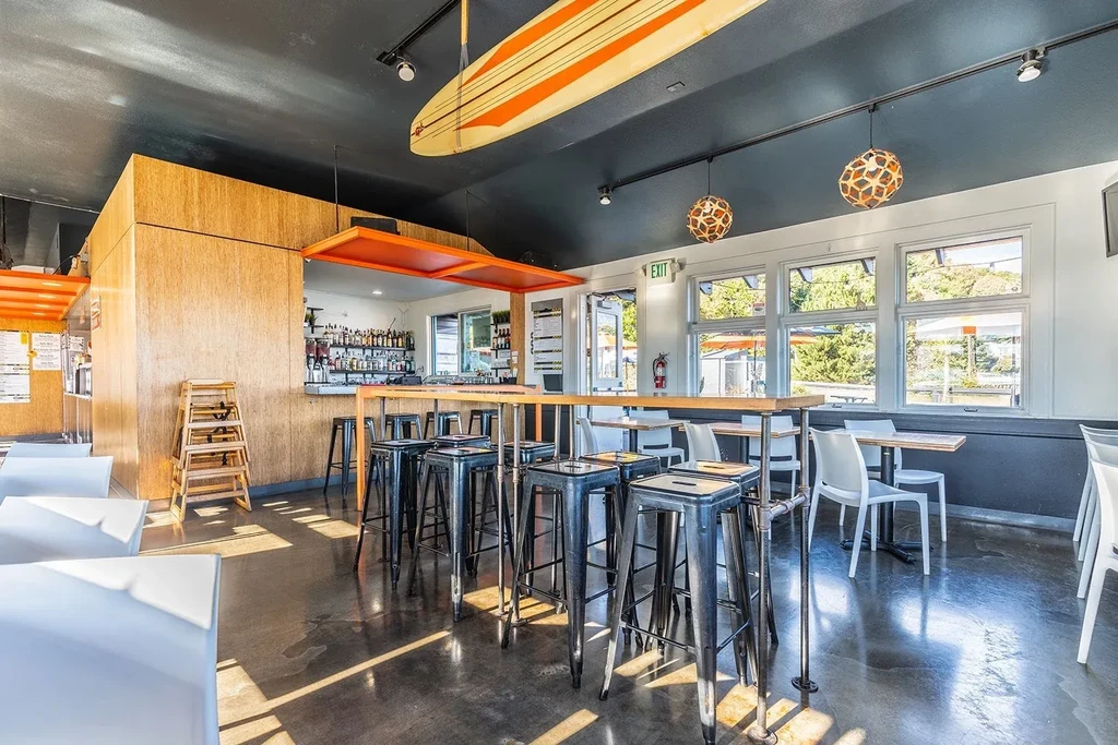 Sunlit modern restaurant with high-top bar, liquor bottles, and surfboard.