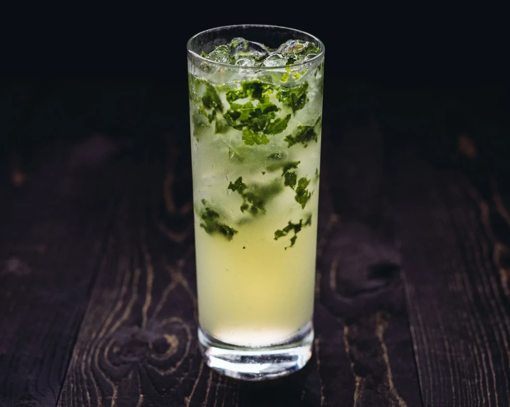 Tall glass with yellow-green drink, crushed ice, mint, on dark wood.