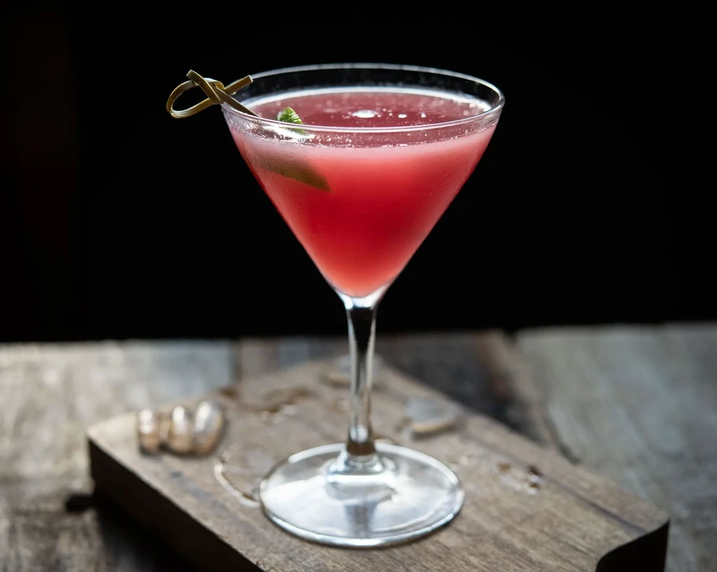Red-pink cocktail in martini glass with leaf on rustic wood.