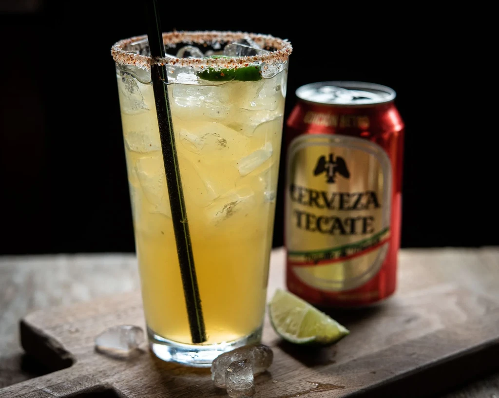 Icy cocktail with chili-salt rim, lime, and Tecate beer can on wood.