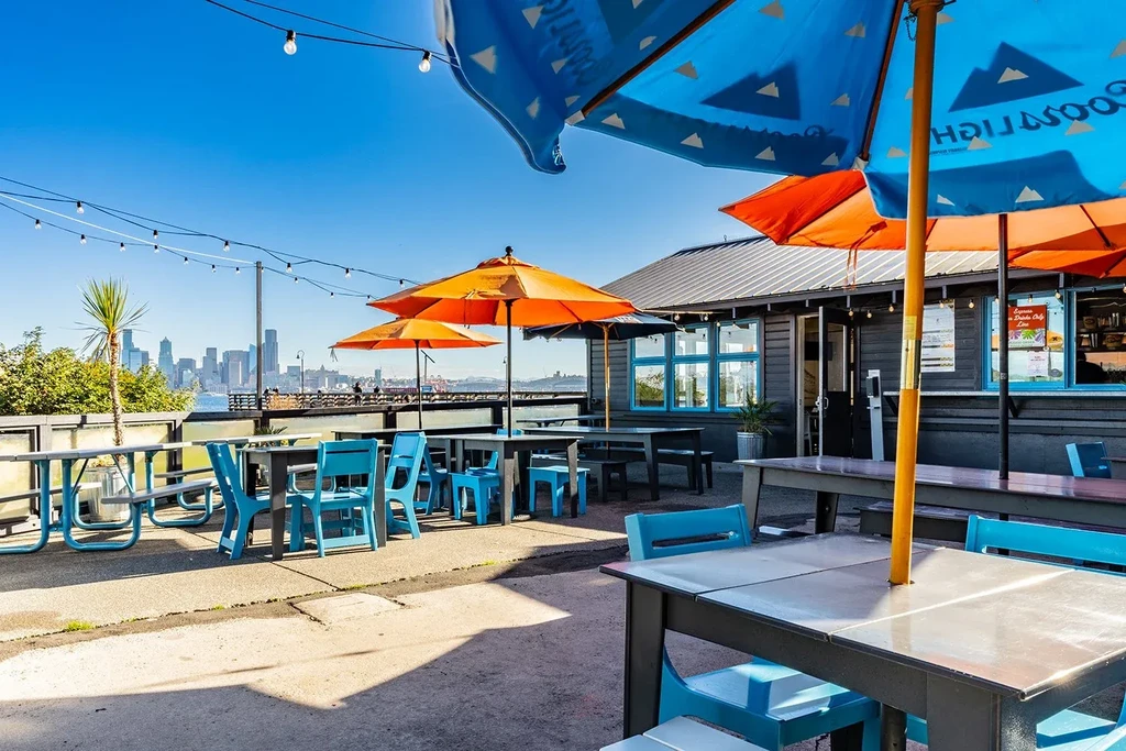 Sunny restaurant patio with blue chairs, umbrellas, string lights, city view.