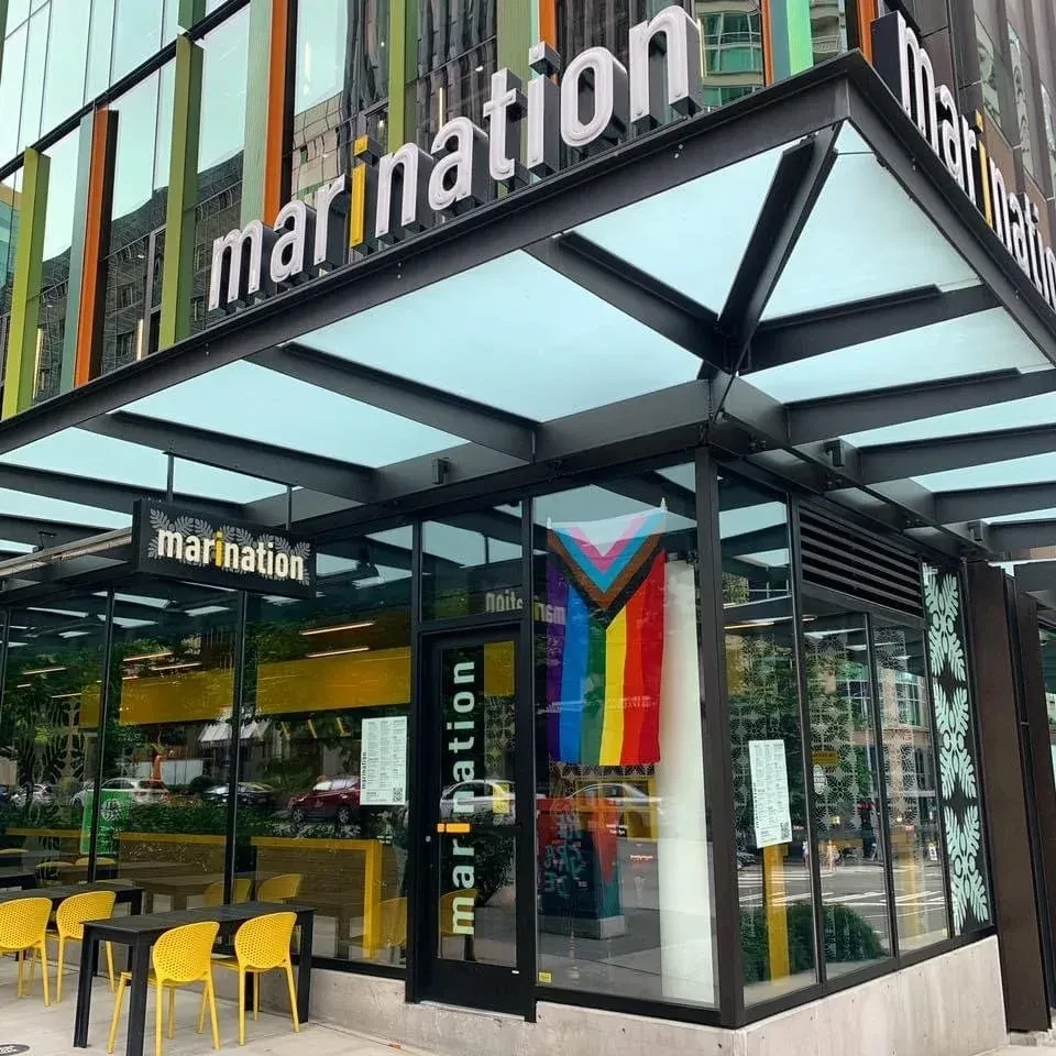 Contemporary "marinaton" restaurant exterior features yellow seating and a Progress Pride flag.