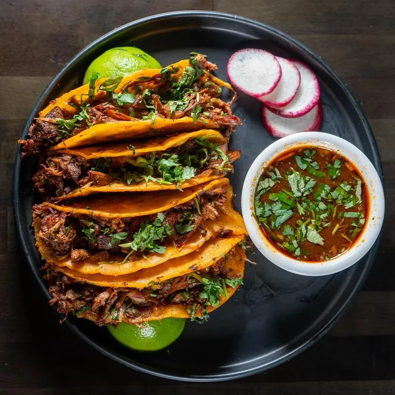 Birria tacos with consommé, lime, and radish, top-down view.