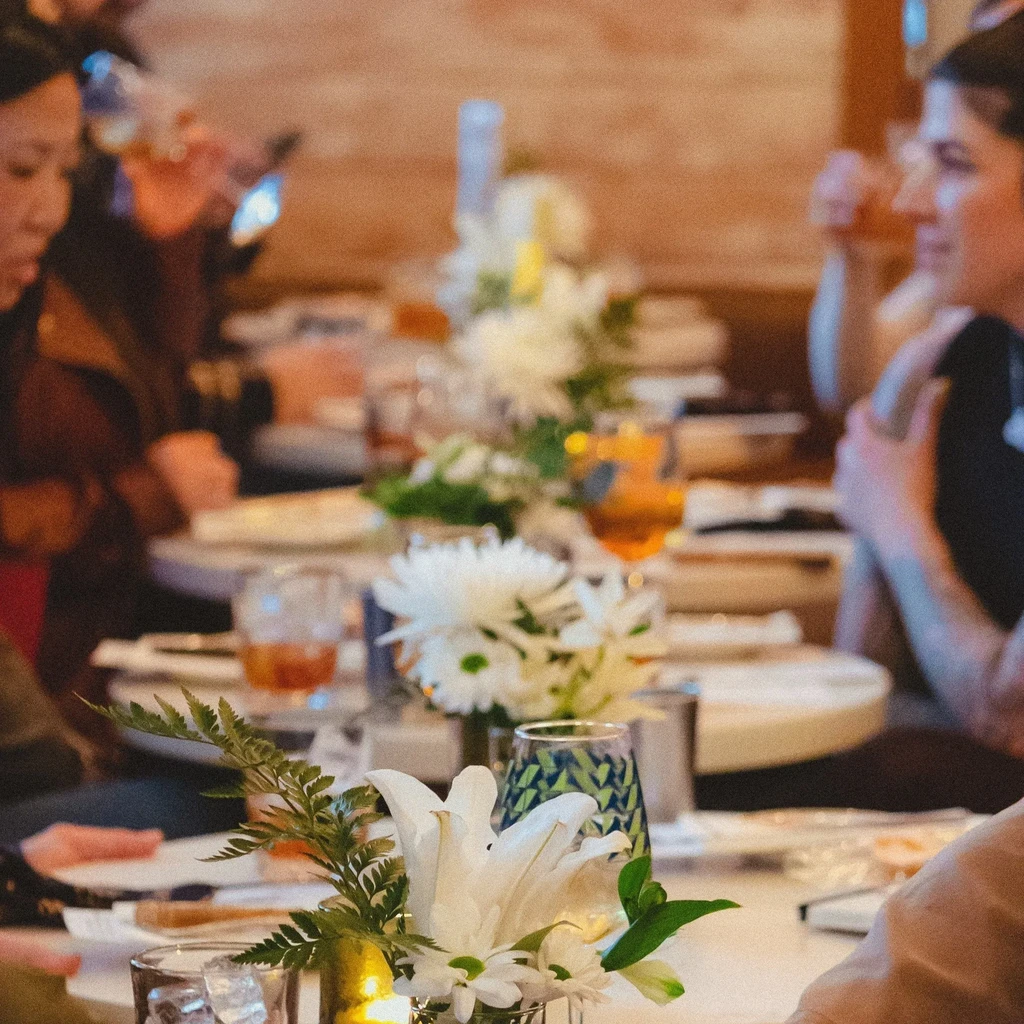 Warm-lit dining table with white flowers, drinks, blurred background.