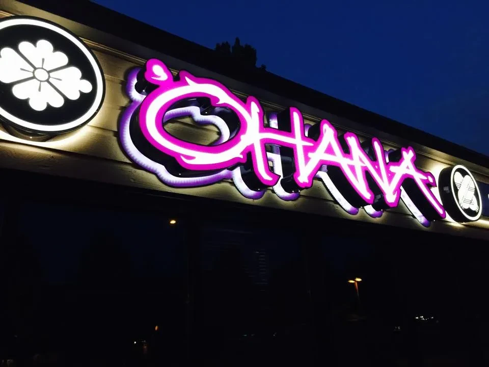 Vibrant pink and white neon 'OHANA' sign with emblems at night.