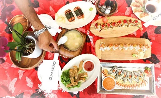 Overhead flat-lay of diverse Asian food on a red table, hand reaching.