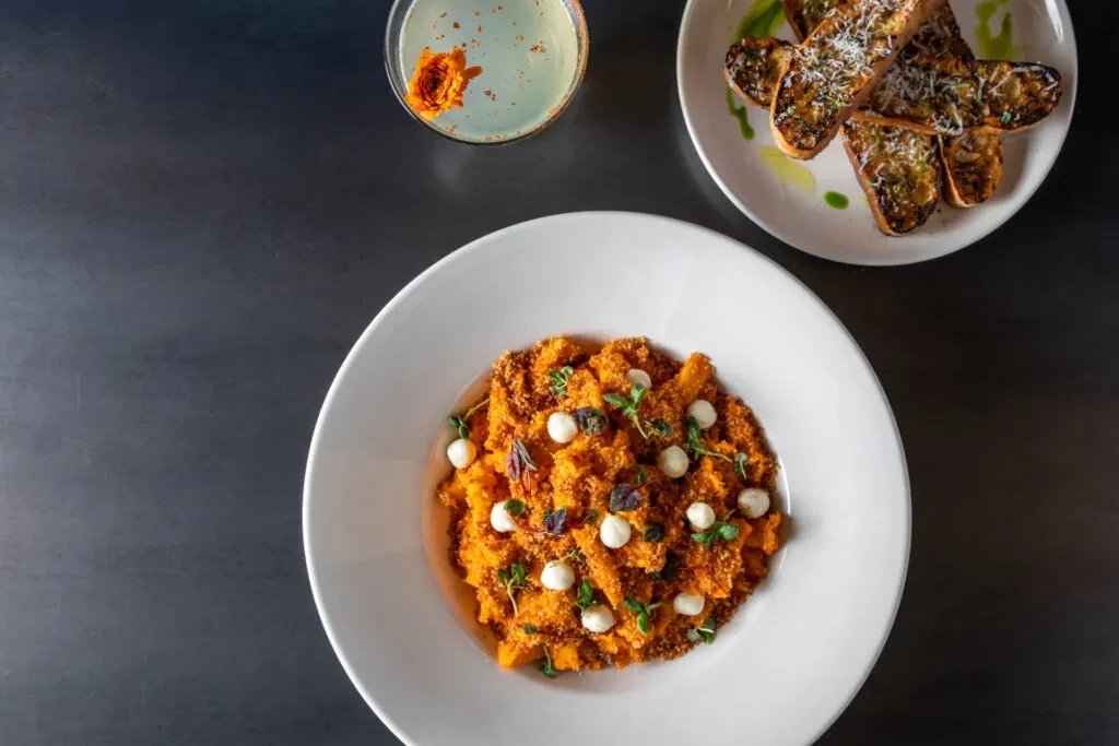 Overhead shot of orange pasta, bruschetta, and a cocktail on a dark table.