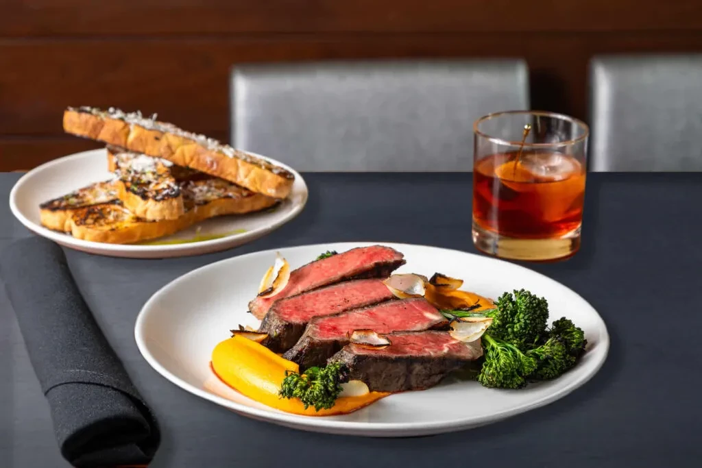 Medium-rare steak, toasted bread, and amber cocktail on a restaurant table.