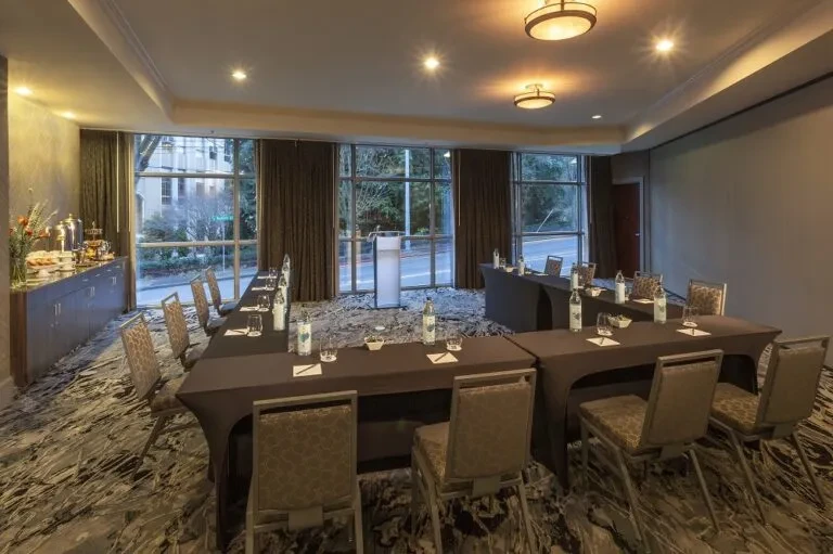 Empty conference room with U-shaped tables, podium, buffet, and windows.