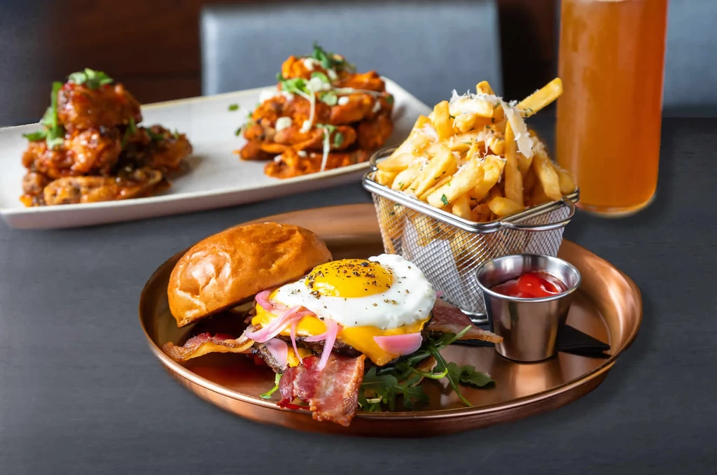 Burger with egg and bacon, fries, wings, drink on dark table.