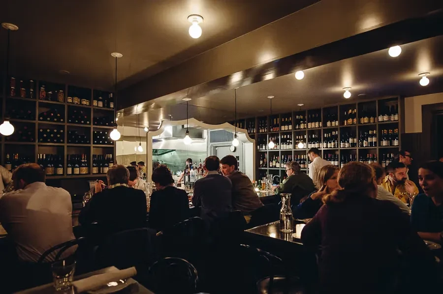 Dimly lit restaurant bar with wine bottles, open kitchen, and many patrons.