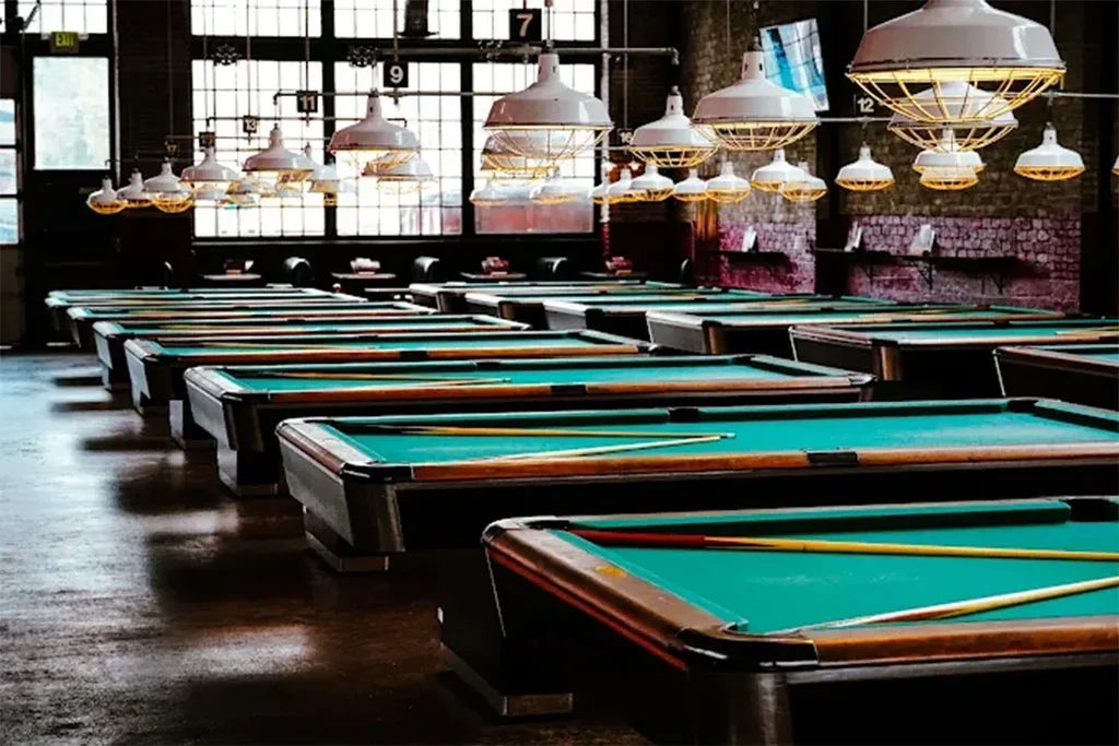 Numerous green felt pool tables arranged in rows in an industrial hall.