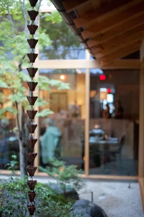 Bronze rain chain foregrounds green foliage and blurred building interior.