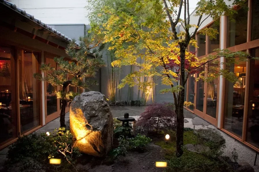 Illuminated Japanese courtyard garden with rock, trees, and restaurant beyond.
