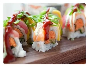 Sushi rolls with fish, avocado, herbs, and sauce on wooden board.