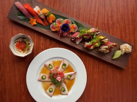 Artfully plated Japanese sushi, sashimi, noodles, and crudo on a wooden table.