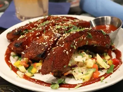 Plate of glazed ribs with sesame, herbs, colorful salad, and dipping sauce.