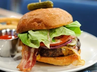 Gourmet burger with cheese, bacon, lettuce, and a skewered pickle.