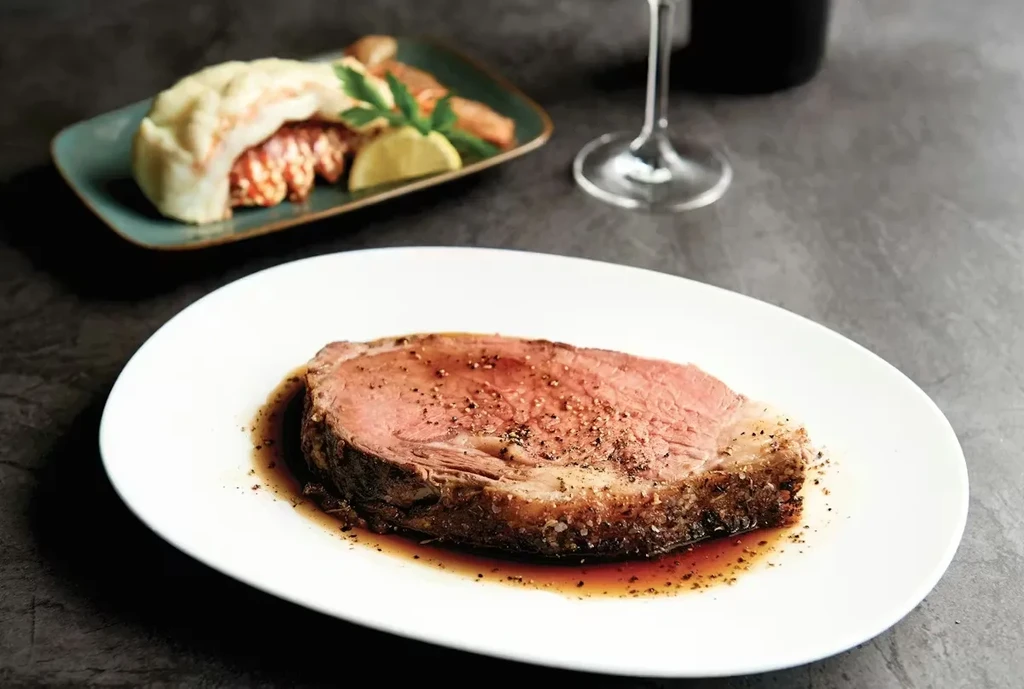 Thick slice of medium-rare prime rib with lobster tail and wine.