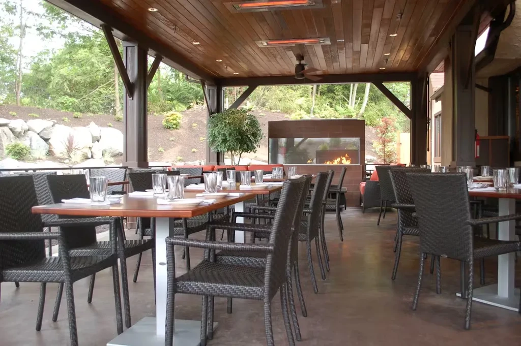 Empty outdoor patio with wood ceiling, lit fireplace, set tables.