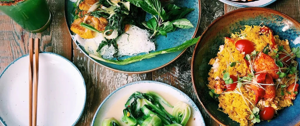 Top-down view: colorful seafood, noodles, vegetables, and a drink on wood.