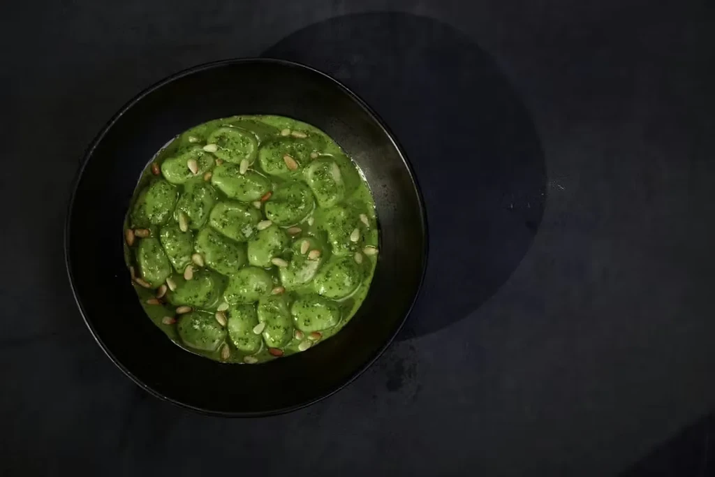 Dark bowl of gnocchi coated in green sauce, garnished with pine nuts.