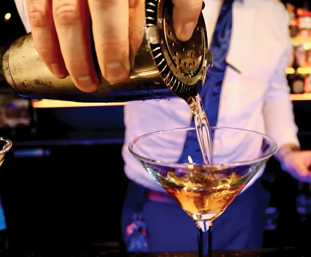 Professional bartender pours golden cocktail from shaker into martini glass.