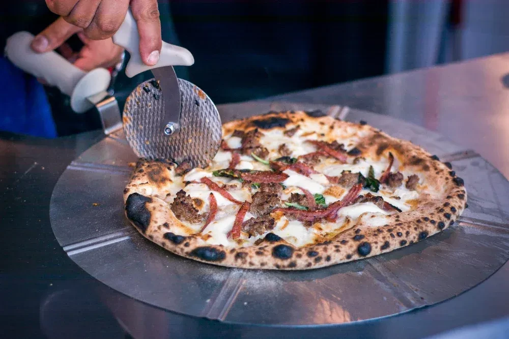 Hands slicing a freshly baked pizza with charred crust and toppings.