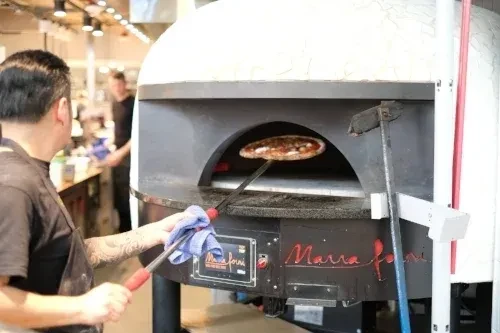 Chef handles pizza in a large, white wood-fired oven.