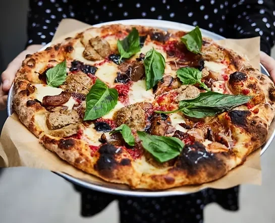 Person holding a rustic pizza with melted cheese, sausage, and basil.