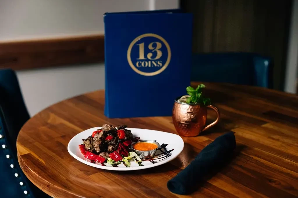 Restaurant table with plated meat dish, Moscow Mule, and "13 COINS" menu.