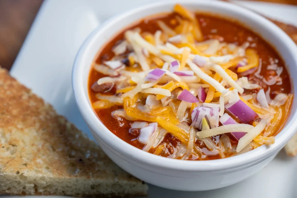 White bowl of chili topped with cheese, red onions, and toast.