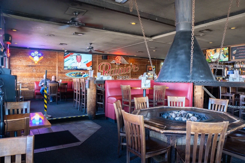 Rustic bar interior with fire pit table, red booths, and neon signs.