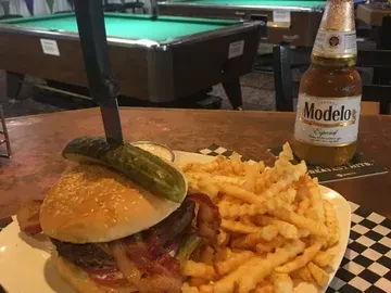 Bacon burger with crinkle fries, pickle skewered by knife, and Modelo beer.