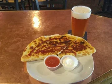 Quesadilla wedges with red drizzle, dipping sauces, and beer on copper table.