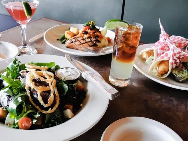 Restaurant table displaying plates of steak, fish, tacos, and cocktails.