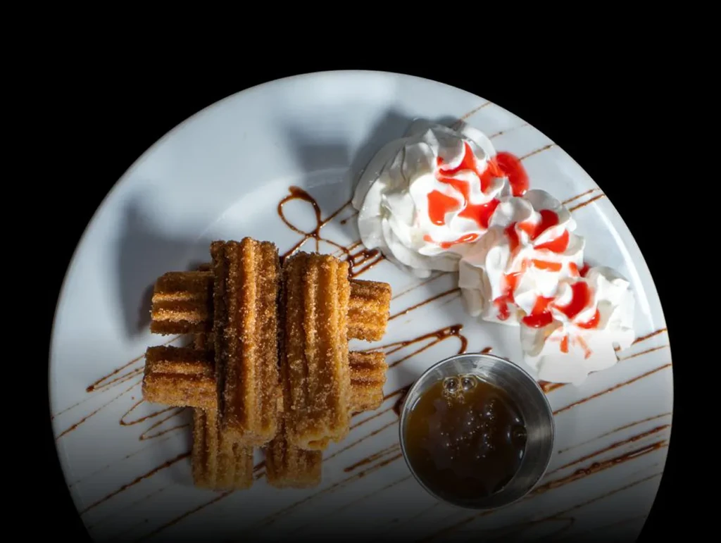White plate of sugar-coated churros, whipped cream, and dark dipping sauce.