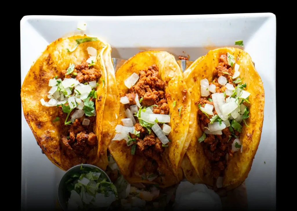 Three tacos with reddish meat, onions, cilantro, on a white plate.