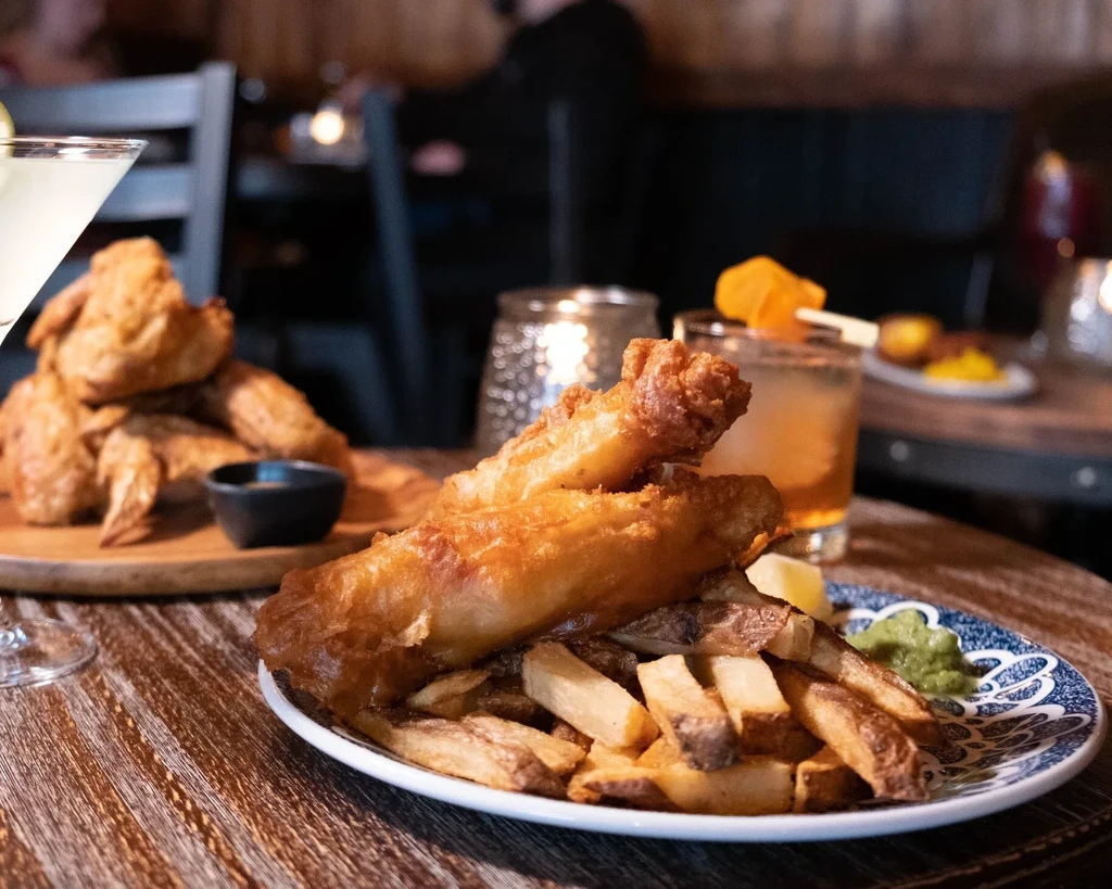 Fish and chips, peas, lemon, cocktails, and chicken wings on a table.