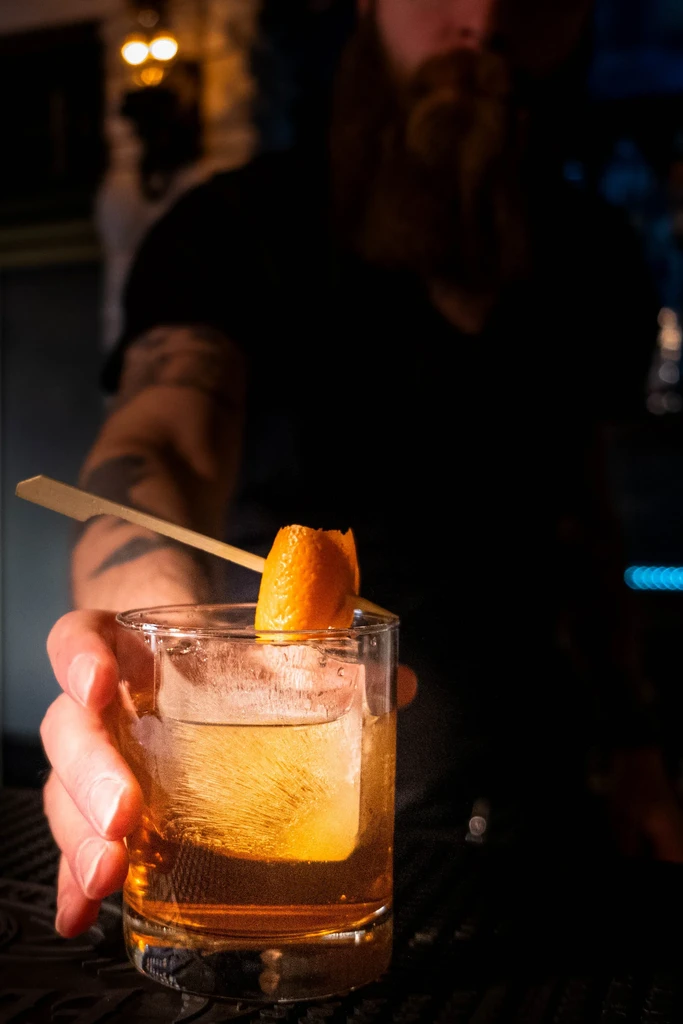 Hand holds amber cocktail with custom ice, orange peel, blurred bartender.