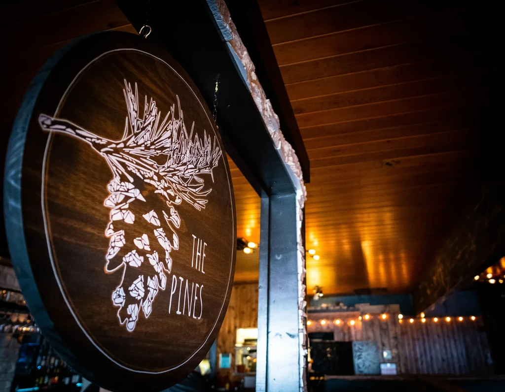 Circular wooden sign "THE PINES" with glowing pine motif in rustic interior.