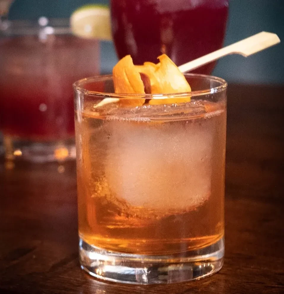 Amber cocktail with spherical ice, orange peel, wooden pick.