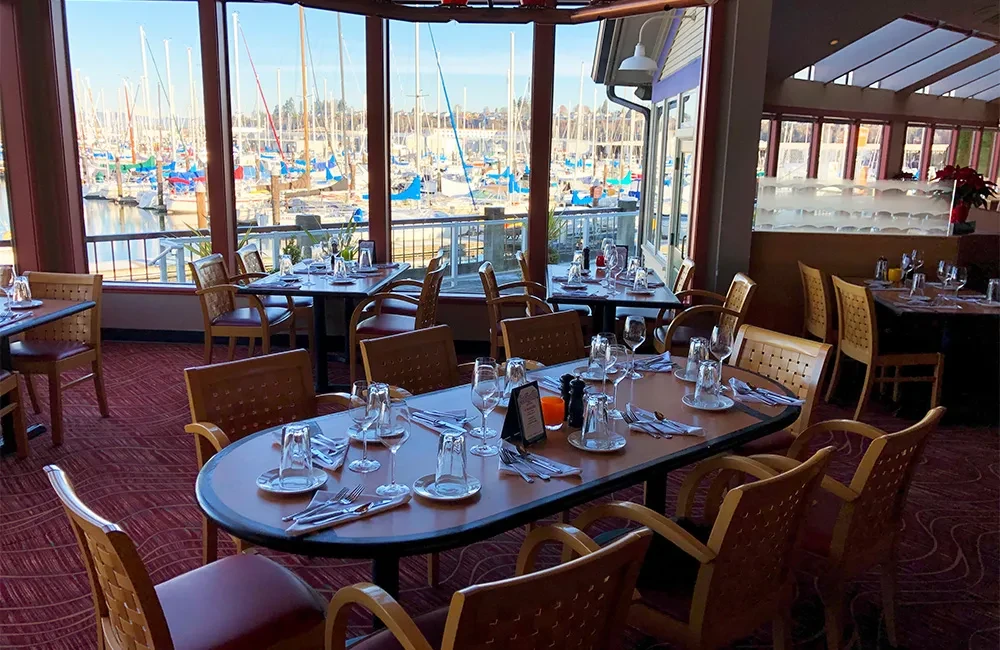 Empty restaurant dining room with set tables and sunny marina sailboat view.