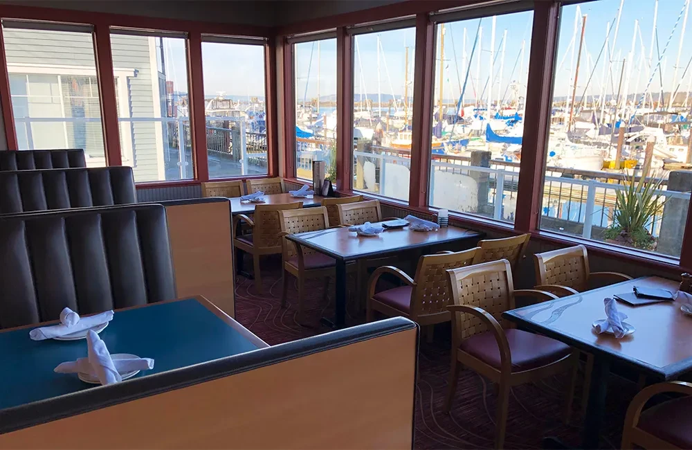 Empty restaurant dining area with sunny marina view and sailboats.